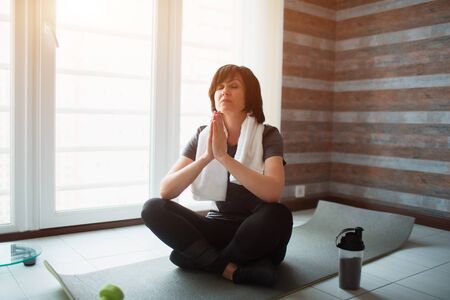 Adult fit slim woman has workout at home. Senior model sit with crossed legs and meditating or praying. Stretching hands holding together. Take care about body and soul.の写真素材
