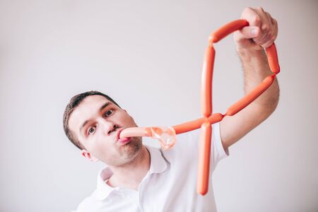 Young man in white shirt isolated over background. Playful positive guy eating sausages. Posing on camera with funny facial expression.の写真素材