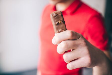 Young man in red shirt isolated over white background. Close up hand holding swet snack. Tasty delicious chocolate bar.の写真素材