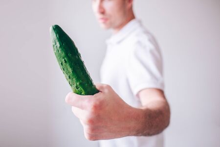 Young man in white shirt isolated over background. Guy look at long cucumber in hand. Cut view blurred defocused background.の写真素材