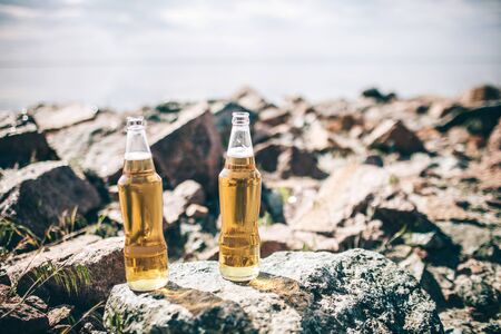 close-up two beer bottles stand on stones near the water in the sunshine against the skyの写真素材