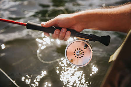 Young fisherman fishing on lake or river. Close up and cut view of spinning reel on rod. Guy hold it in one hand during fishing time. Sunny day at river or lake waterの写真素材