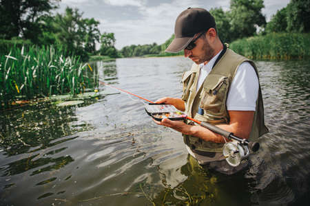Young fisherman fishing on lake or river. Concentrated busy guy hold opened box with plastic lures...