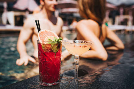 Young hot couple resting at swim-pool. Front view of two glasses of tasty delicious cocktails.