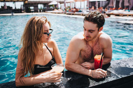 Young hot couple resting at swim-pool. Sexy gorgeous man and woman stand in water and look at each...