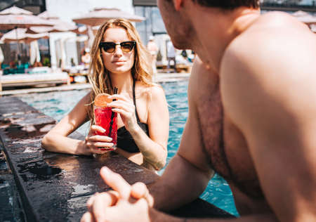Young hot couple resting at swim-pool. View of sexy gorgeous woman looking at man and drinking cocktails.の写真素材