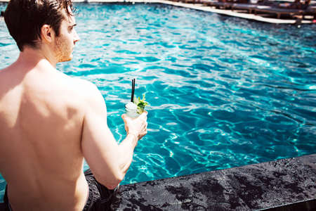 Young hot man resting at swim-pool. Cut back view of strong powerful guy holding cocktail in hand and enjoy spending time at water.の写真素材