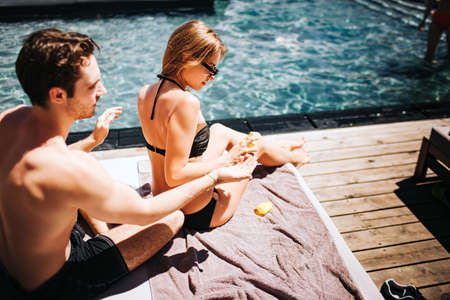 Young hot couple resting at swim-pool. Guy sit behind girl and using sun protection cream for back...