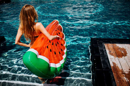 Young hot woman resting at swim-pool. Back view of female model going down in water steps with swim ring in hand.の写真素材