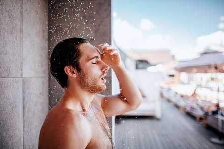 Young man taking shower outside. Side view portrait of guy without shirt standing under shower while water falling down. Enjoy water procedure.の写真素材
