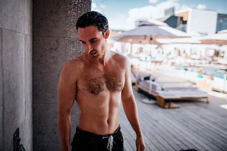 Young man taking shower outside. Strong muscled and powerful guy having water procedures. Dropping down. Washing body and head. Alone outside.の写真素材