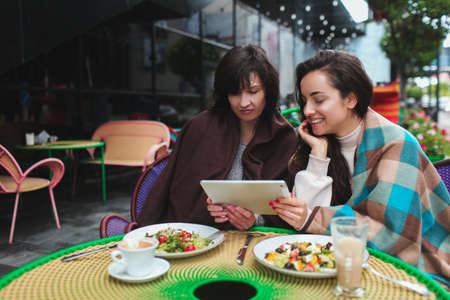 Mature mother and her young daughter sit together in cafe or restaurant. Hold and look together at tablet in hands. Modern technologies. Warm generation friendship. Nice meal in restaurant.の写真素材