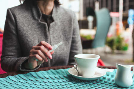 Cut view of business woman sit alone at cafes table holding electronic cigarette in hand. Cup of tea stand on top. Small coffee break or lunch time. Bad habit of smoking. Doesnt care about health.の写真素材