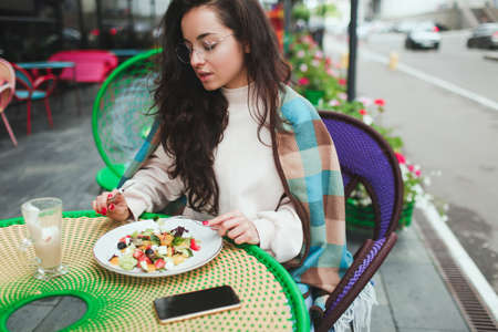 Young woman sit alone in cafe or restaurant. Serious busy girl eating salad outside during lunchtime. Sitting alone keeping smartphone besides her.の写真素材
