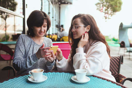 Mature mother and her young daughter sit together in cafe or restaurant. Girl shows her mother something on yellow smartphone. Watching together and having fun. Using modern technologies.の写真素材