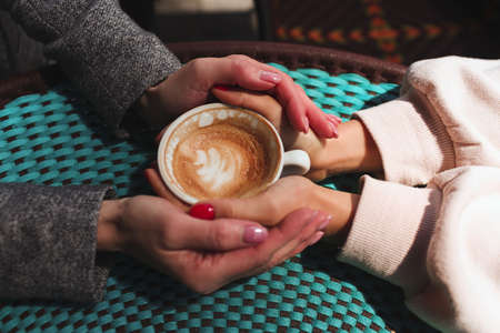 Mature mother and her young daughter sit together in cafe or restaurant. Up view of female hands holding one cup of coffee together. Love and support. Family connection.の写真素材