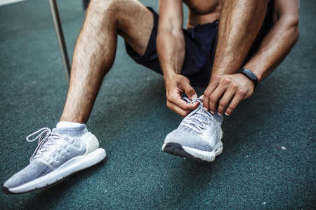 Young man exercising outside. Cut view of sportsman sitting on fround and tie shoe laces. Preparing...