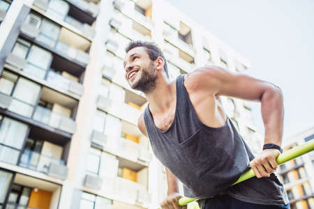 Young man exercising outside. Cheerful positive sportsman holding hands on sporty bar and keep his...