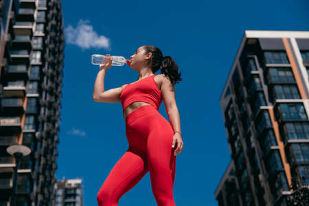 Pretty sporty girl with brown ponytail wearing red leggins and top drinking water from plastic bottle. Resting athletic woman. Blurred city background. Low angle shot.の写真素材