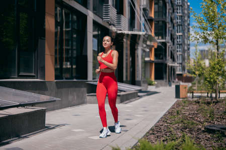 Sporty pretty woman jogging on tiled pavement in urban area. Fit girl with dark ponytail wearing in red sport top, leggins and white sheakers. Apartment block and trees in background.の写真素材