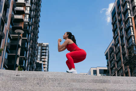 Side view of athletic pretty girl with dark ponytail squating on top of the stairs. Sporty woman...