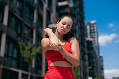 Front view of beautiful young woman in red bra feeling pain in her elbow, hand. during sport workout outdoors. Close-up view. Blurred apartment blocks on background.の写真素材