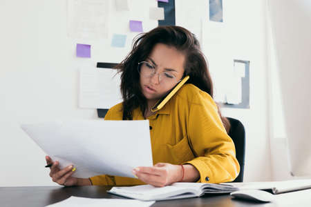 Confused young woman reading documents and discussing on the phone. Startup bussines concept. Woman in yellow clothes and in round eyewear talking by her phone. Work in office or from home.の写真素材