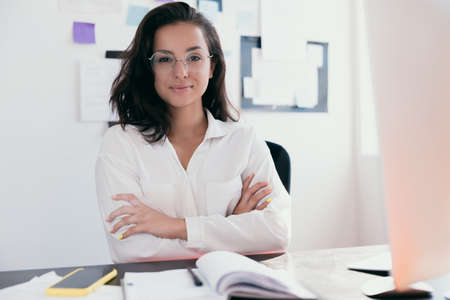 onfident and successful female leader keeping arms crossed and looking at camera with smile. Woman with brown hair in round glasses wears white shirt. Office work or work at home concept.の写真素材