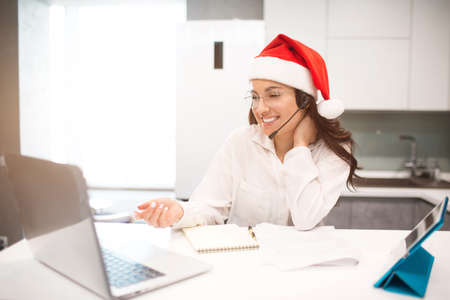 Merry New Year and Merry Christmas. Beautiful black-haired woman works from home. An employee sits in the kitchen and has a lot of work on a laptop and tablet and has video conferencing and meetings.の写真素材