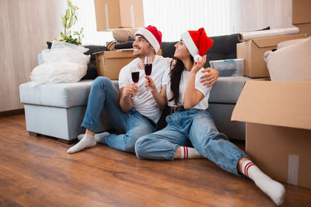 Newlywed couple celebrate Christmas or New Year in their new apartment. Young happy man and woman drinking wine, celebrating moving to new home and sitting among boxes.の写真素材