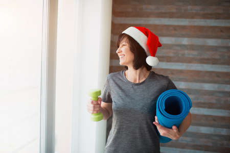 Adult fit slim woman has workout at home. New year or Christmas time period. Well-built adult senior woman resting after workout in room. Hold green dumbbell and blue yoga mat.の写真素材