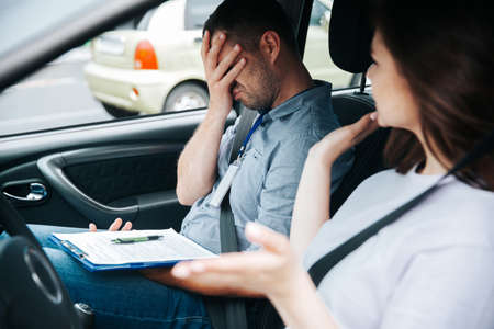 Upset male driving examiner in gray shirt and jeans makes facepalm gesture with one hand and holds clipboard with another. Young woman in white shirt fails drivers license exam.の写真素材