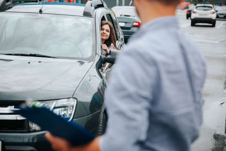 Blurred man with clipboard in hands turns around and looks at attractive young woman in grey car. Caucasian woman smiles and invites him to join the trip.の写真素材