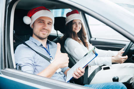 Young female driving student and male instructor in santa claus hats sit in a car together. Thumb up gesture. View through car window. Merry Christmas and Happy New Year. Driving courses concept.の写真素材