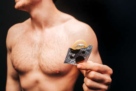 Cropped image of man holding opened package with condom in hand. Obscure face of sexual muscular guy with naked sportive torso. Concept of sex protection. Isolated on dark background. Studio shootの写真素材