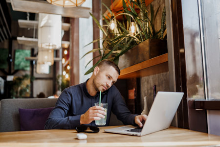 Young businessman is talking on phone while working on laptop in restaurant. Remote work.の写真素材