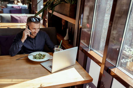 businessman is talking on phone, while having lunch and working on laptop in restaurant.の写真素材