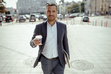 Young businessman holding his laptop while drinking coffee. Walking on the street.の写真素材