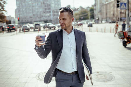 Young businessman holding his laptop while drinking coffee. Standing on the street.の写真素材