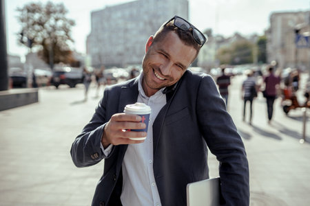 Young businessman holding his laptop while using his phone and drinking coffee. Standing on the street.の写真素材