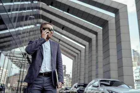 A handsome young businessman holding his laptop while using his phone and standing on the streetの写真素材