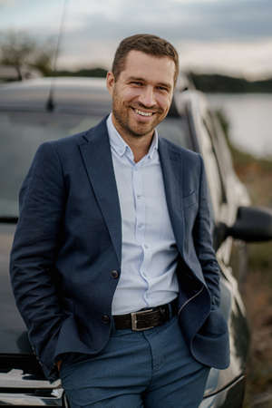 Young smiling man with black classic car wearing black suit. Man is standing near his car outdoors.の写真素材