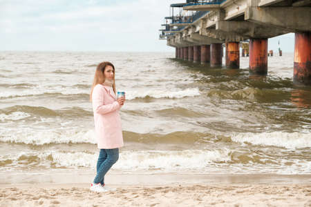 the girl is standing on the seashore. wearing a pink coat. drinks coffee..girl hiker standing near a tent and holding a cup lake and mountains in the backgroundの写真素材