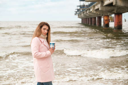 A young beautiful woman in a pink coat stands on the sea or ocean shore with a cup of coffee and enjoys the view. brunette on the seashore drinking coffee. glass with coffee.の写真素材