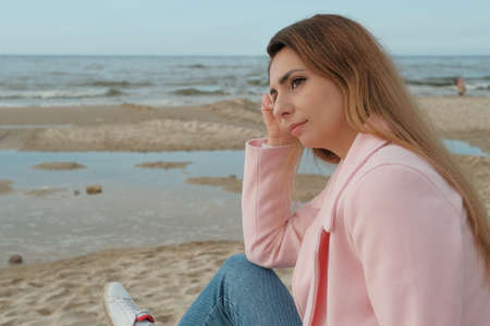 A young beautiful woman in a pink coat stands on the sea or ocean shore with a cup of coffee and enjoys the view. brunette on the seashore drinking coffee. glass with coffee.の写真素材
