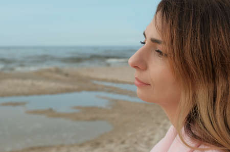 A young beautiful woman in a pink coat stands on the sea or ocean shore with a cup of coffee and enjoys the view. brunette on the seashore drinking coffee. glass with coffee.の写真素材
