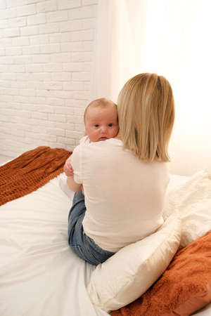 mom holds a newborn baby in her arms. in the background the sun shines from the window.