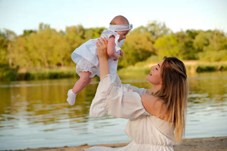 a beautiful girl in a white dress with a newborn baby is sitting on the beach, on the sand.の写真素材