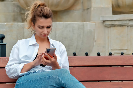 a beautiful girl sits on a bench and holds a phone in her hands. the girl orders food through the phone. the girl is talking on the phone laughing, happy, surprised.の写真素材