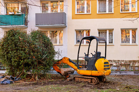 Yellow mini excavator in front of a multi-storey building next to a green bush. High quality photoの写真素材